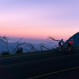 Two women cycling up a mountain with a backdrop of a pink and blue sky